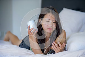 Young beautiful Asian Chinese woman with earpiece listening to music smiling happy lying on bed using internet on mobile phone