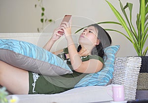 Young beautiful and relaxed Asian Chinese woman lying at home living room sofa couch using internet on mobile phone happy and comf