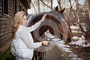 Young beautiful rider woman blonde with long hair in white clothes posing with brown horse