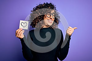 Young beautiful romantic arab woman wearing glasses holding reminder paper with heart pointing and showing with thumb up to the