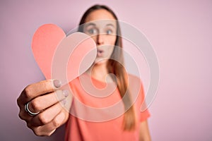 Young beautiful romantic woman holding paper heart shape over isolated pink background scared in shock with a surprise face,