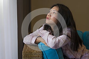 Young beautiful sad and depressed Asian Chinese woman in pajamas at home sitting by living room couch feeling unhappy and confused