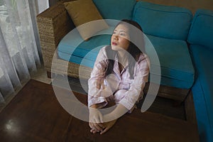 Young beautiful sad and depressed Asian Korean woman in pajamas at home sitting by living room couch feeling unhappy and confused