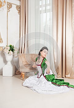 Young beautiful sad woman in rococo style medieval dress sitting on the floor near armchair