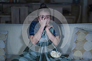 Young beautiful scared and frightened Asian Japanese woman watching horror scary movie or thriller eating popcorn in fear face