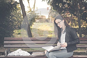 Young beautiful school or college girl with eye glasses sitting on the bench in the park reading the books and study for exam, kno