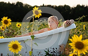 Young beautiful serene blonde sexy woman, with a sponge, taking relaxed a bath in nature in the warm sunset with yellow sunflowers