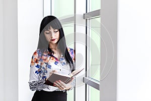 Young Beautiful serious woman standing near the window, writing in notebook, looking to the notebook