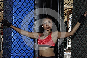 Beautiful and Asian fighter woman in fighting gloves and sport clothes inside MMA cage posing cool