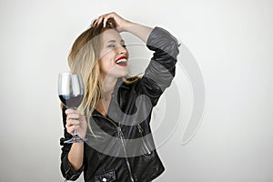 Young beautiful sexy blonde woman wearing black leather jacket drinks wine from glass laughing on isolated white