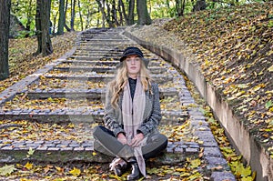 Young beautiful girl model posing in the autumn park among fallen yellow leaves on the old stairs in a hat, coat, jeans and b