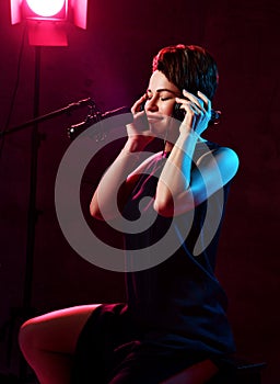 Young beautiful short haired brunette woman singer in black dress sitting in studio in headphones and listening to music