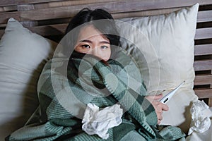 Young beautiful sick and exhausted Asian Chinese woman suffering cold and flu holding thermometer having temperature lying on bed