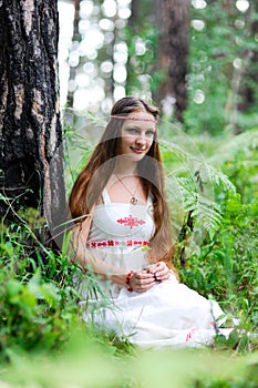 A young beautiful Slavic girl with long hair and Slavic ethnic attire sits in a summer forest and holds a berry in her hands