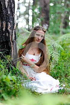 A young beautiful Slavic girl with long hair and Slavic ethnic attire sits in a summer forest and holds a berry in her hands