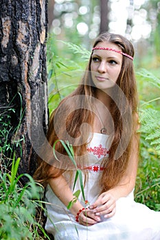 A young beautiful Slavic girl with long hair and Slavic ethnic attire sits in a summer forest and holds a berry in her hands