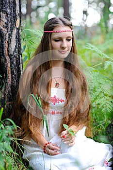 A young beautiful Slavic girl with long hair and Slavic ethnic attire sits in a summer forest and holds a berry in her hands