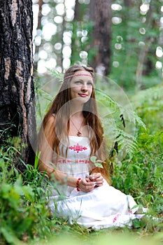 A young beautiful Slavic girl with long hair and Slavic ethnic attire sits in a summer forest and holds a berry in her hands