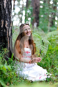 A young beautiful Slavic girl with long hair and Slavic ethnic attire sits in a summer forest and holds a berry in her hands