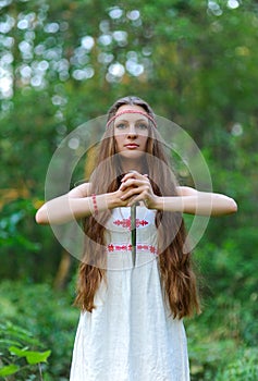 A young beautiful Slavic girl with long hair and Slavic ethnic dress stands in a summer forest with a ritual dagger in her hands