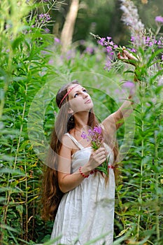 A young beautiful Slavic girl with long hair and a Slavic ethnic dress stands among the tall undergrowth in the summer forest and