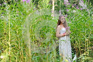 A young beautiful Slavic girl with long hair and a Slavic ethnic dress stands among the tall undergrowth in the summer forest and