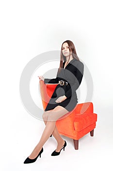 A young, beautiful, slender girl in a black dress and black shoes stands next to a red armchair on a white background. Isolated. L