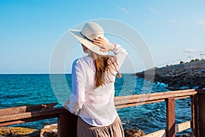 Young beautiful slender woman in sunhat with long hair in boho style clothes at the shore looking and the sea clear blue sky