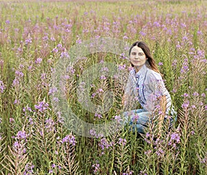 Young beautiful smiling blond woman in purple shirt sitting in the meadow among flowers of fireweed