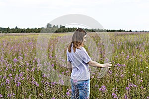 Young beautiful smiling blond woman in purple shirt walking in meadow among flowers of fireweed and touching flower