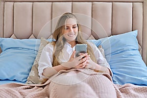 Young beautiful smiling blonde with smartphone lying in bed at home