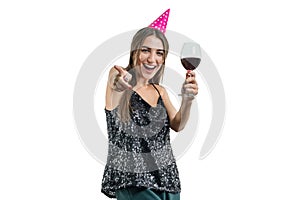 Young beautiful smiling brunette woman with glass of red wine and wearing party hat points her finger at you, isolated on white