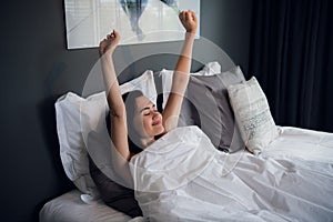 Young beautiful smiling and brunette woman lying in bed trying to wake up early morning stretching hands after sleep