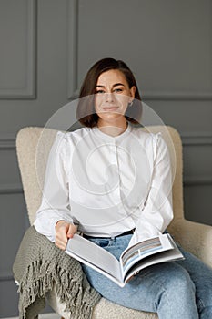 Young beautiful smiling business woman wearing a white shirt and jeans with a magazine in her hands, sitting in a beige chair with
