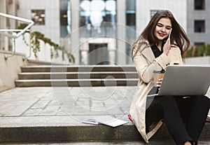 Young beautiful smiling businesswoman works in her laptop drinks coffee sitting on stairs near office center in downtown, looks