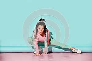 Young beautiful smiling girl with modern dreadlocks hairstyle in sportswear and sneakers sitting on floor and doing stretching