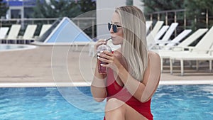 A young beautiful smiling girl is resting by the pool drinking cherry cocktail