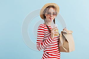 Young beautiful smiling girl with two braids in straw hat and red sunglasses drinking orange juice and holding paper bag