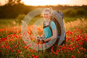 Young beautiful smiling woman with backpack and walking sticks stands on field of poppies