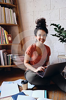 Young beautiful smiling woman with dark curly hair sitting on floor with laptop on knees happily looking aside with