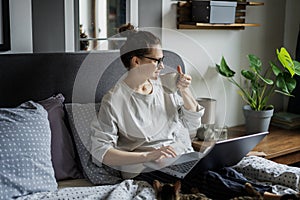 Young beautiful smiling woman drinking morning coffee and using laptop while lying in bed at home