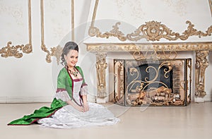 Young beautiful smiling woman in green rococo style medieval dress sitting near fireplace