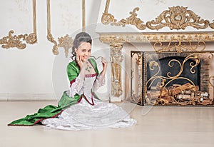 Young beautiful smiling woman in green rococo style medieval dress sitting near fireplace