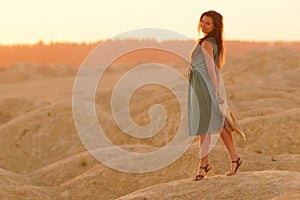 Young beautiful smiling woman with long hair in long blue dress with straw hat in hand on sand at sunrise in desert