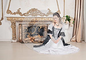 Young beautiful smiling woman in rococo style medieval dress sitting on the floor near fireplace