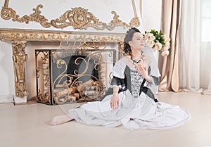 Young beautiful smiling woman in rococo style medieval dress sitting on the floor near fireplace