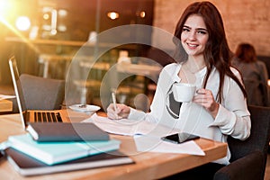 Young beautiful smiling woman working outside office in her laptop taking notes in her notebook while drinking hot coffee in the