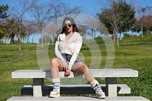 Young and beautiful Spanish brunette woman with sunglasses sitting on a park bench. The girl is casually dressed and makes