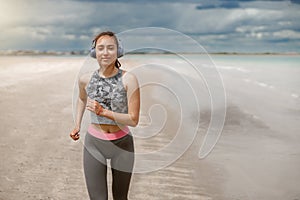 Young beautiful sportive woman in headphones running over seaside on morning at beach