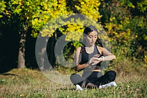 Young beautiful sports girl asian woman meditating in park, sitting lotus pose practicing yoga mat outdoors looking smart watch at